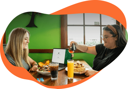 Two woman at a table next to a window with an open laptop, two meals, and two beverages. 