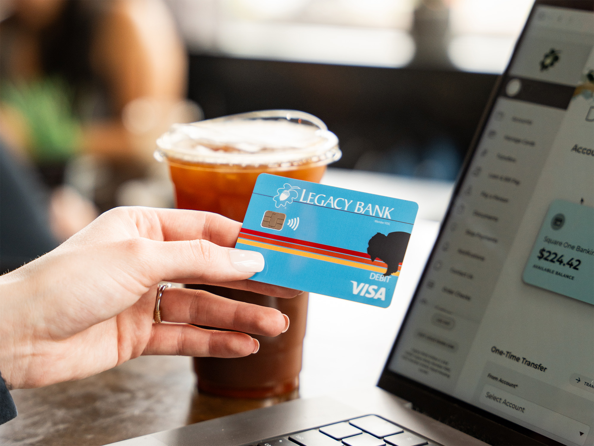 Blue Legacy Bank Check card with a laptop in the background of a coffee shop. 